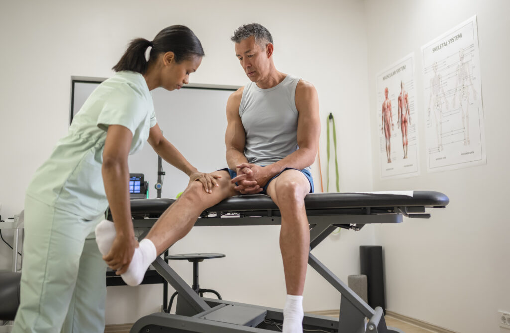 Man in consultation for joint injury or inflammation. Female Chiropractor or physiotherapist consults with patient about rehabilitation and recovery from muscle injury at rehabilitation clinic. Health and care concept.